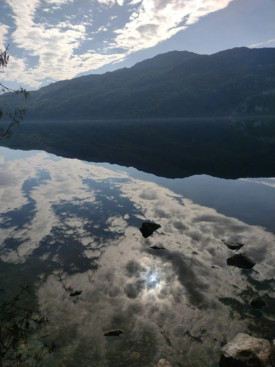 Loch Ness on a still day, with clouds, sky and the hillside on the far shore reflected almost perfectly in the water and a few dark rocks in the shallows