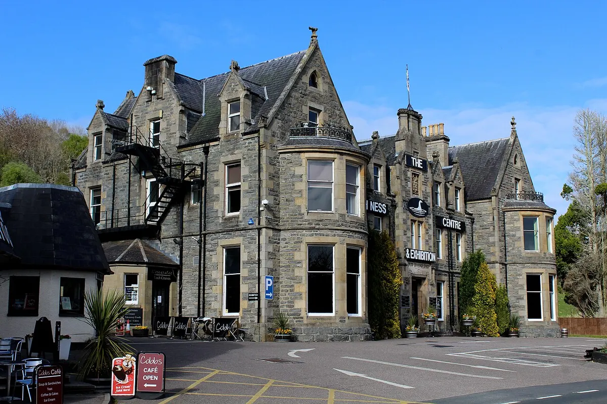 A large grey stone building with gables and bay windows, signed “The Loch Ness Centre & Exhibition,” with a café and parking area in front