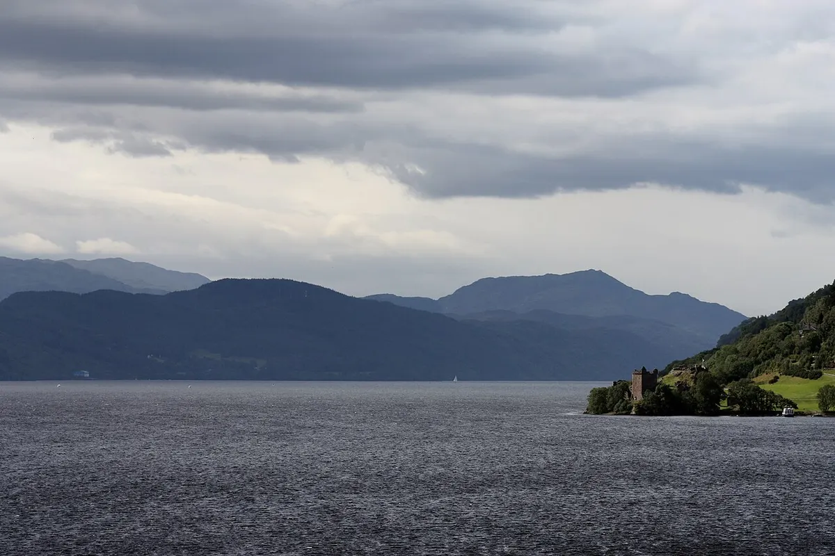 Loch Ness under heavy grey cloud, with the ruins of Urquhart Castle on a green headland at the right and hills along the far shore