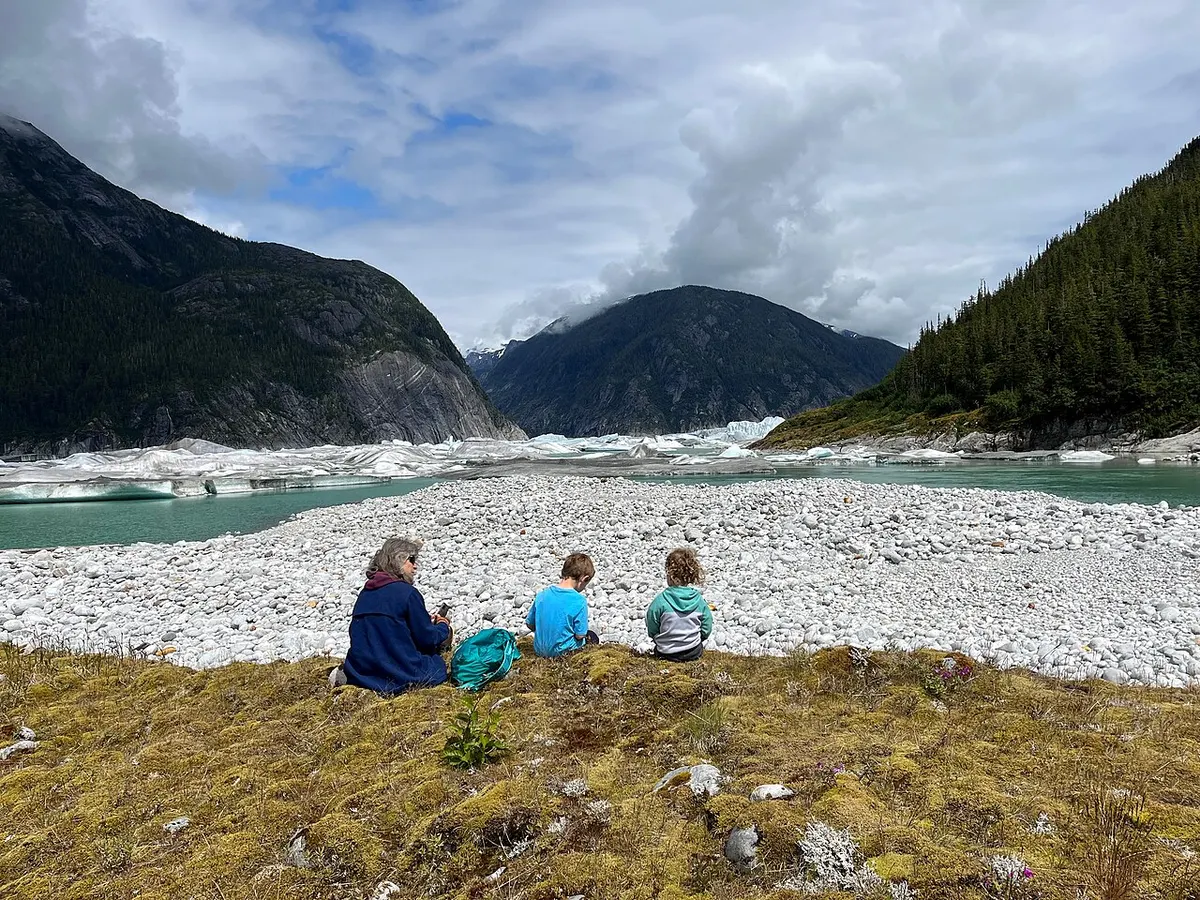 Three people sit on a mossy bank above a wide gravel flat and an iceberg-filled lagoon below the Baird Glacier, with steep forested mountains on either side