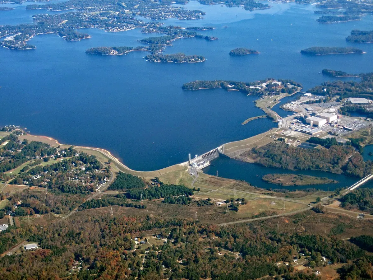 Aerial view of Lake Norman’s wide blue water and wooded islands, with the long earthen Cowans Ford Dam and its hydroelectric station in the foreground