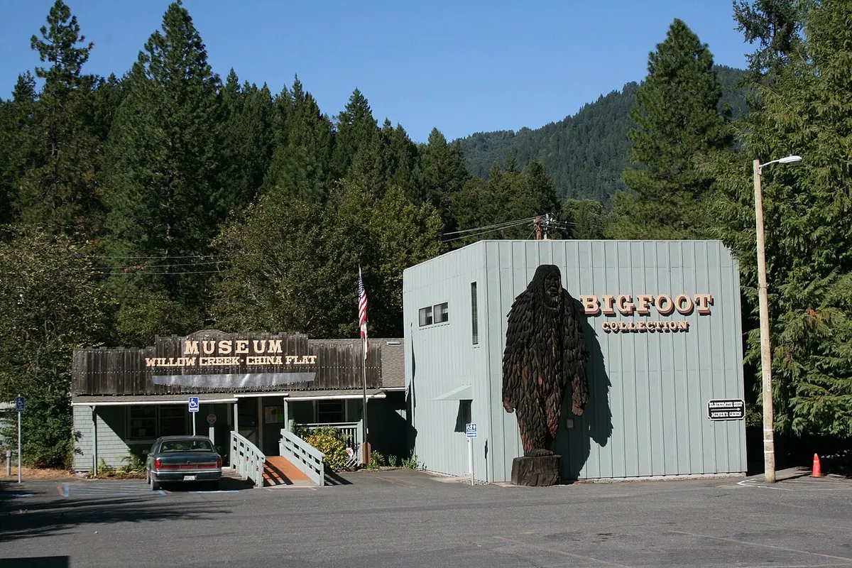 A tall carved wooden Bigfoot statue stands outside the Bigfoot Collection building at the Willow Creek–China Flat Museum, with forested hills behind