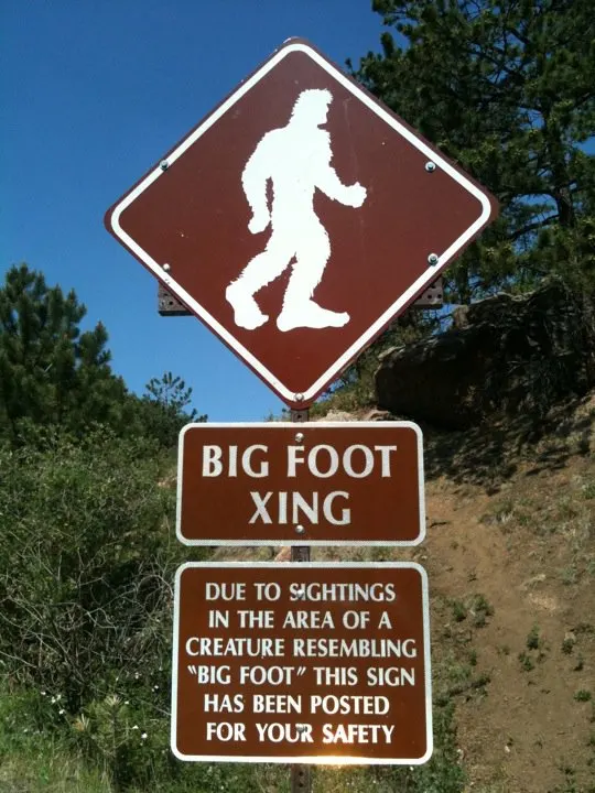 A brown diamond sign with a white Bigfoot silhouette above a “Big Foot Xing” plate and a warning notice on Colorado’s Pikes Peak Highway