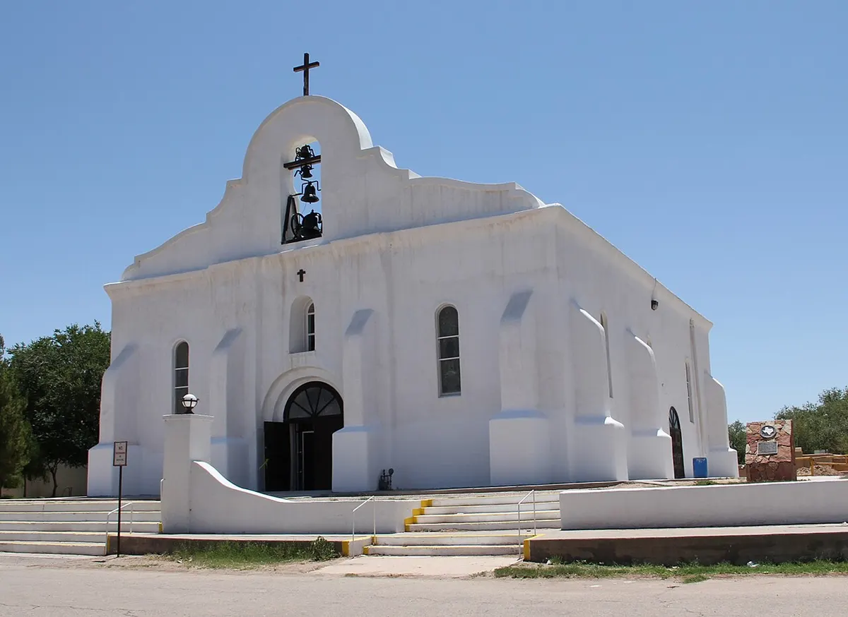 A whitewashed adobe-style chapel with a bell gable and three bells under a clear blue sky in San Elizario, Texas
