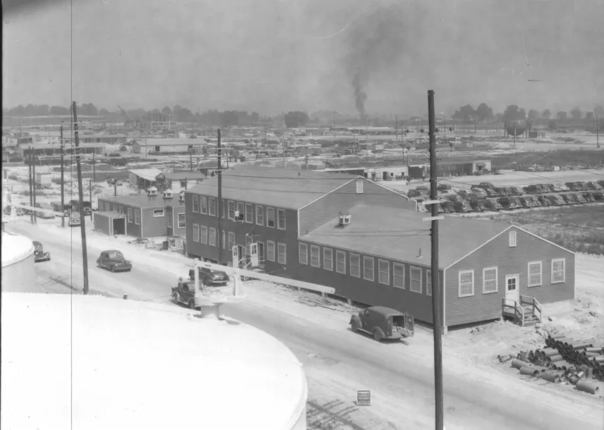 Black-and-white photograph of a two-story wartime laboratory building beside a road lined with 1940s cars, with the sprawling plant behind